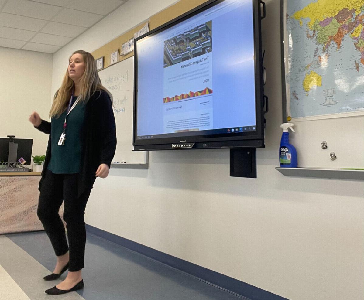 A student teacher stands in the front of a classroom in front of a large screen